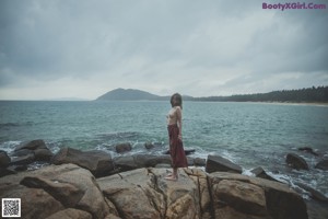 A naked woman standing on a rock by the ocean.